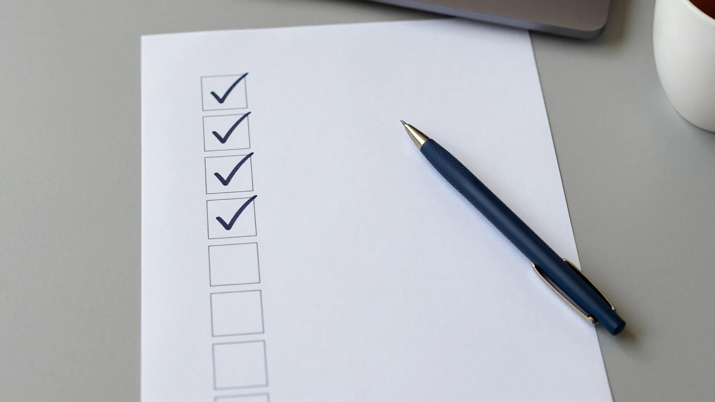 Illustration: a sheet of paper with a column of boxes, four ticked and the rest empty, a navy pen resting on the sheet and a cup of tea at the edge.